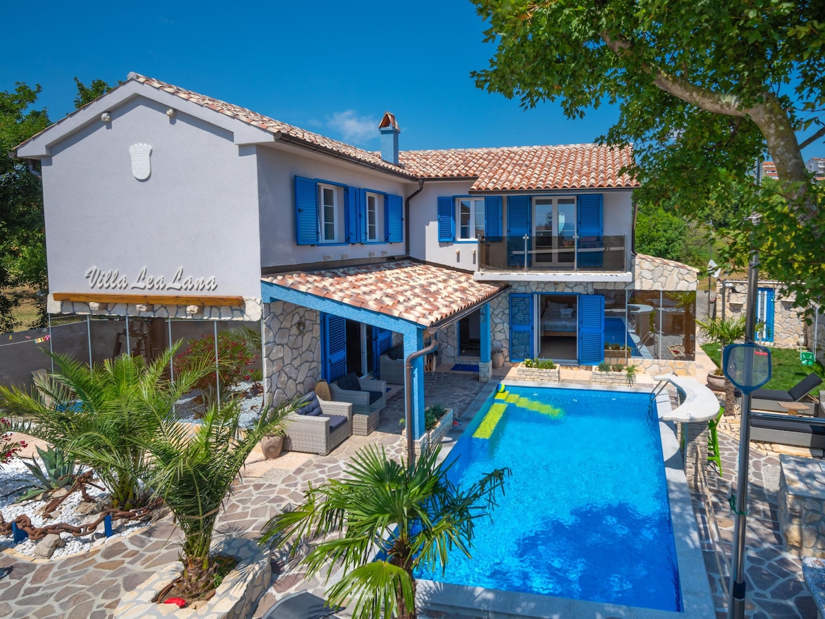 The villa features a charming two-story design with blue shutters and a tiled roof. A clear swimming pool is situated in front, surrounded by palm trees and outdoor seating areas. The pathway is bordered by white pebbles, enhancing the sunlit garden ambiance.