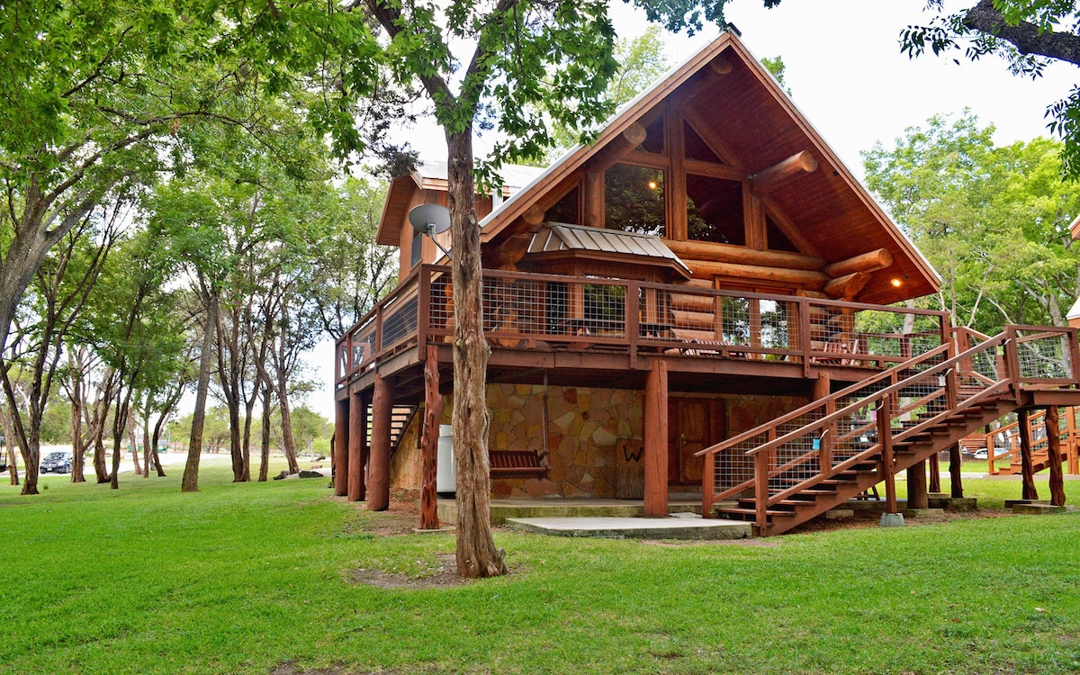 A log home with a spacious deck is showcased amid lush greenery. The structure features large windows and a prominent A-frame roof. Sturdy wooden stairs lead to the entrance, and the surrounding lawn is dotted with trees, enhancing the outdoor experience.