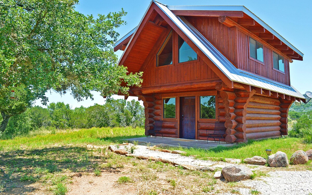 The log cabin exterior features a combination of natural wood and an angled roofline. Large windows enhance the view of the surrounding greenery. A spacious porch welcomes visitors, complemented by landscaped areas and large stones.