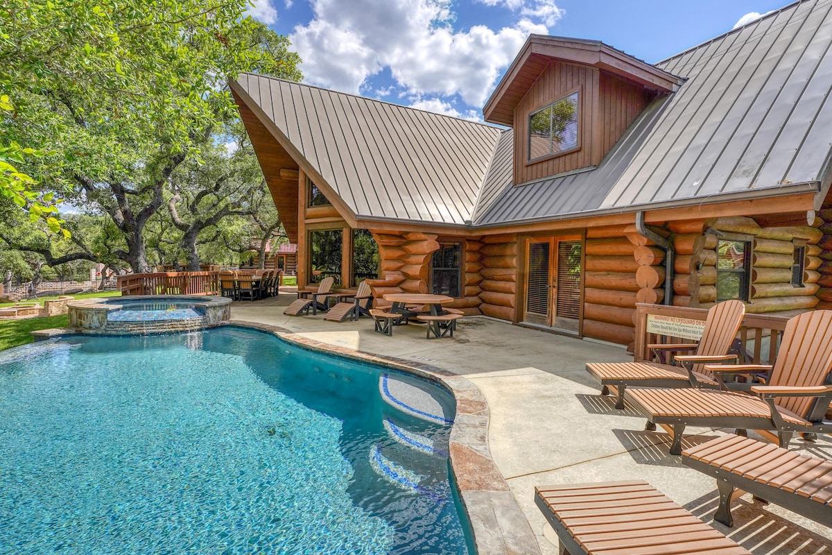 A spacious outdoor area features a crystal-clear pool, surrounded by comfortable loungers. The log home is characterized by a distinctive angled roof and large windows, offering an unobstructed view of the lush greenery. A hot tub is positioned nearby, inviting relaxation.