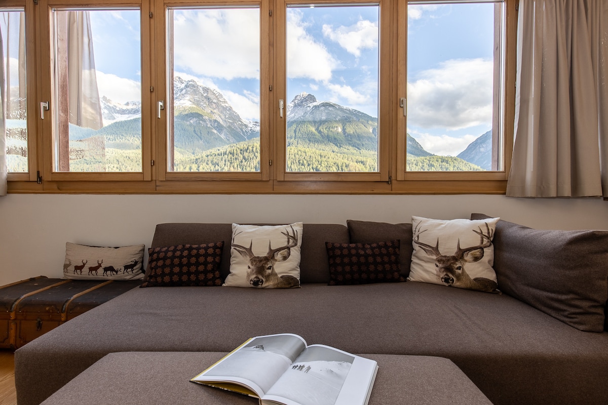 A spacious seating area is highlighted by large windows, offering panoramic views of the surrounding mountains. The couch is adorned with decorative pillows featuring deer motifs. A coffee table holds an open magazine, inviting relaxation and enjoyment of the scenic backdrop.