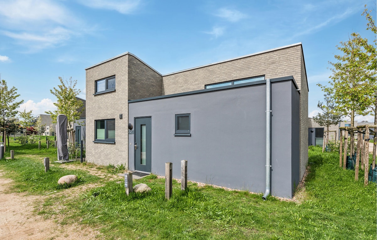 A modern building is showcased, featuring a combination of gray brick and a sleek facade. The exterior design includes large windows and a minimalist entryway. The surrounding landscape is lush with greenery, including small trees and decorative stones.