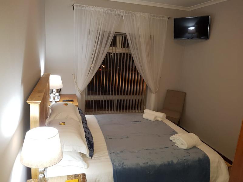 A cozy bedroom is showcased with a spacious bed adorned in neutral linens and a soft blue throw. Nightstands with lamps flank the bed. A soft chair is positioned nearby, while a television is mounted above the window with sheer curtains gently framing the view.