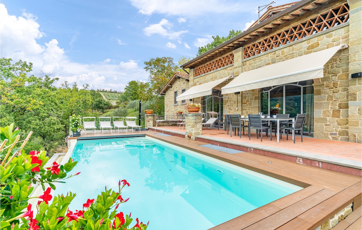 A serene outdoor space features a private pool with a wooden deck, bordered by vibrant plants. Comfortable seating is arranged under large awnings, with a dining table nearby. The rustic stone house and surrounding landscape create an inviting atmosphere for relaxation.