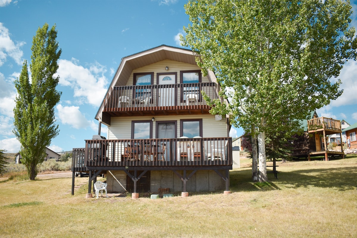 The exterior of the cozy cabin is showcased, featuring a two-story structure with a balcony and deck overlooking the expansive lawn. Comfortable seating arrangements are visible on the deck, surrounded by lush trees and a clear blue sky.