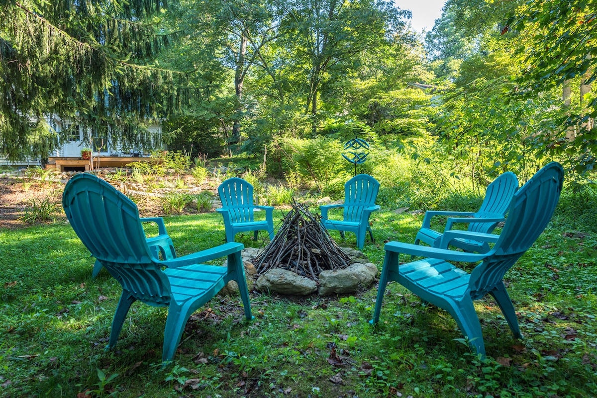 A cozy outdoor gathering area features six turquoise chairs arranged around a central fire pit, with a stack of firewood resting on a stone base. Mature trees provide a lush green backdrop, contributing to a serene and inviting atmosphere.