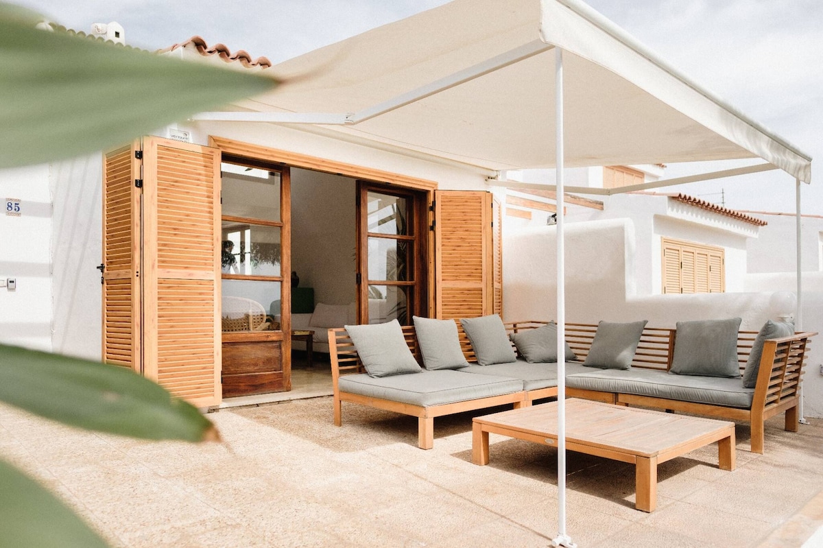 An outdoor seating area features a spacious sofa with soft gray cushions, complemented by a wooden coffee table. The area is shaded by a retractable awning and framed by wooden louvered doors that connect to the interior space.