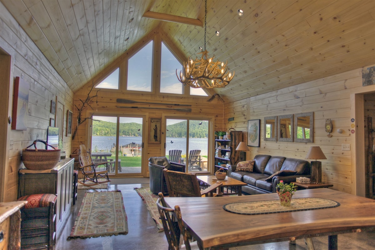 An open-concept living area features vaulted wood ceilings and large windows showcasing views of the lake. Comfortable seating is arranged around a coffee table, with a dining area nearby. A rustic chandelier illuminates the space, enhancing the natural wood decor.