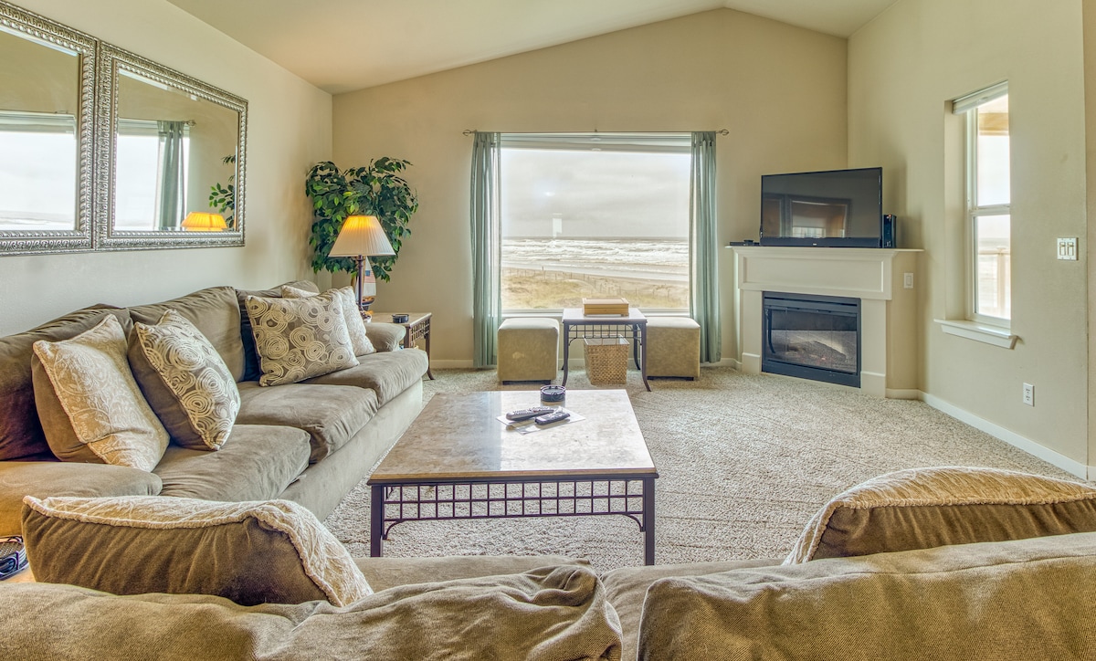 A comfortable living room space features an oversized couch and loveseat arranged around a coffee table. A large window offers views of the ocean, while a flat-screen TV is positioned against the wall. Warm lighting is provided by a floor lamp beside a decorative plant.
