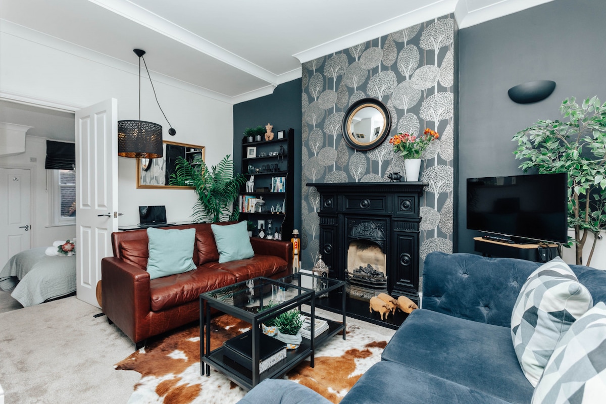 A modern living area features a brown leather sofa adorned with teal cushions and a large blue sofa. A glass coffee table is centered on a patterned rug, with greenery and decorative items. The wall is decorated with a tree-patterned wallpaper, and natural light enters through a nearby doorway.