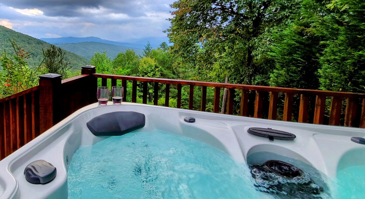 A hot tub is positioned on a raised deck surrounded by natural greenery, offering expansive mountain views in the background. Two glasses of red wine are placed near the edge, inviting relaxation while enjoying the serene landscape.