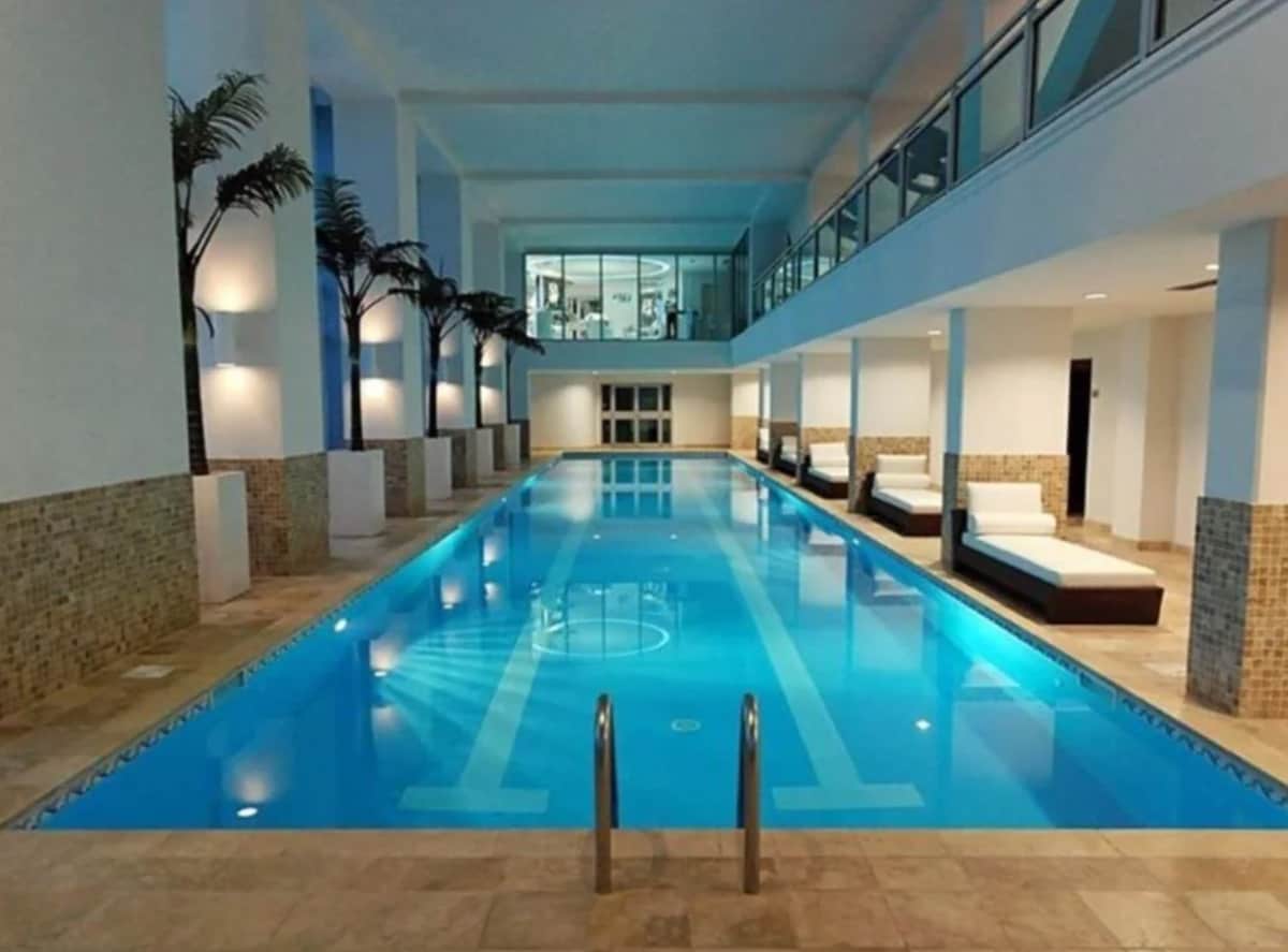 An indoor heated lap pool is framed by light-colored tiles and walls, with palm trees positioned along the edges. Lounge chairs are arranged on both sides, creating a relaxing atmosphere. The pool's clear blue water reflects the overhead lighting, enhancing the serene setting.