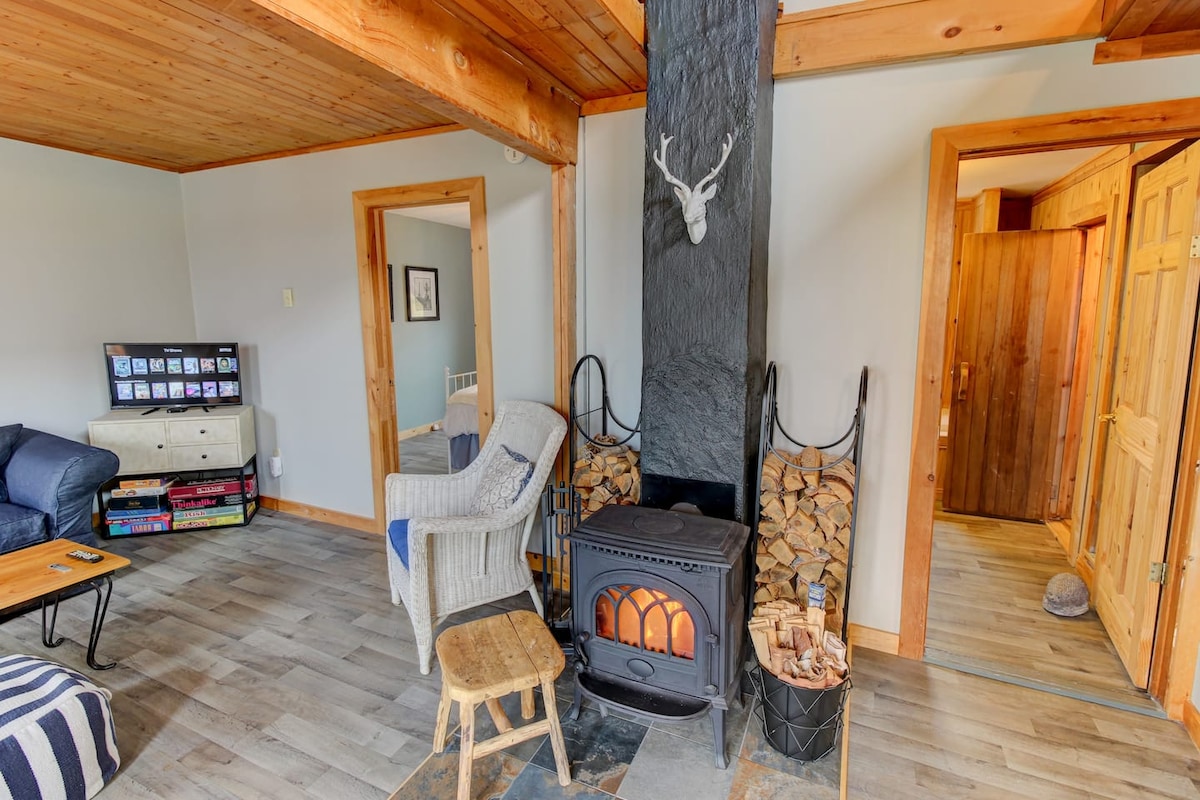 A cozy living area features a wood stove surrounded by stacked logs, complemented by a small wooden stool and a textured chair. Natural wood accents are present throughout the space, with a large couch and a coffee table offering comfort and functionality.