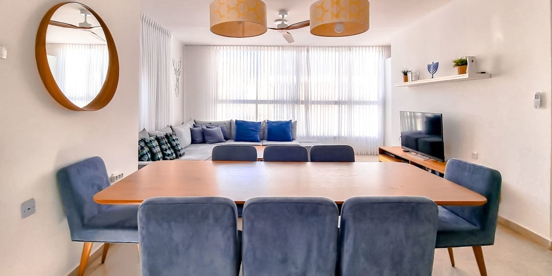 A dining area features a wooden table surrounded by plush blue chairs. A large mirror reflects natural light, while the living space in the background is enhanced by a window with sheer curtains and a cozy seating area with decorative cushions.