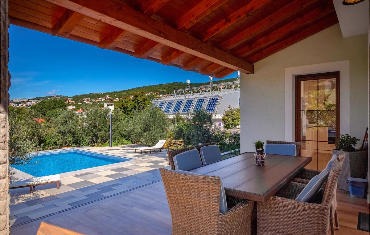 A covered terrace includes a wooden dining table surrounded by woven chairs. The view features a swimming pool surrounded by sun loungers, with lush greenery and distant hills in the background under a clear blue sky.