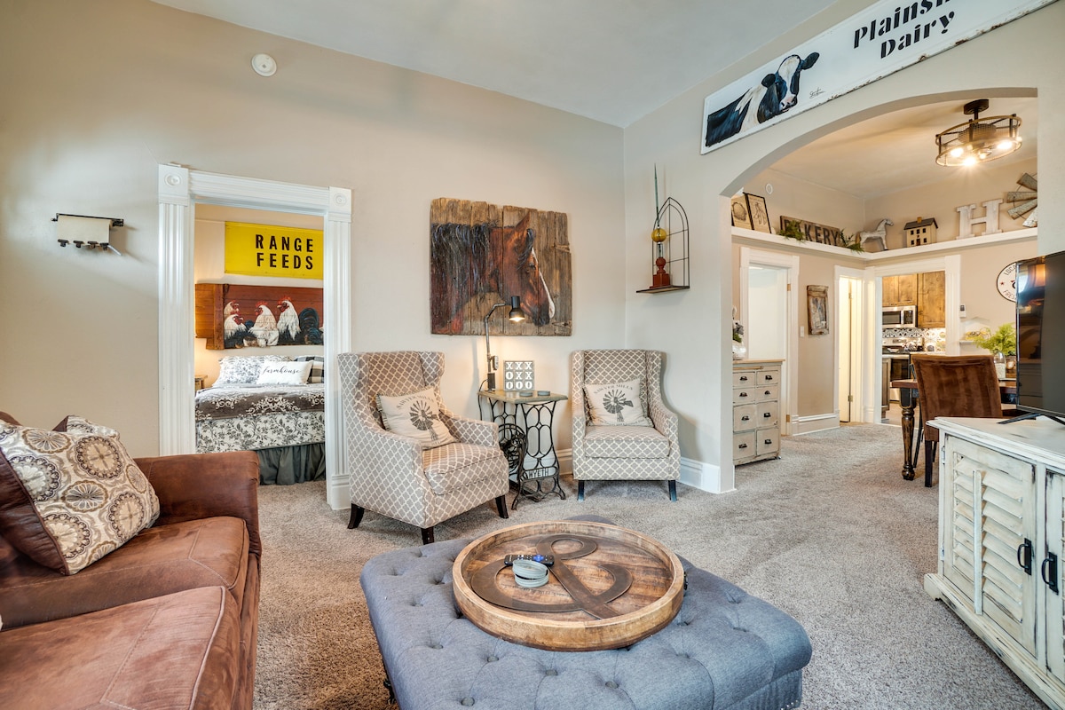 A welcoming living area is presented, featuring a comfortable sofa and armchairs arranged around a coffee table. Decor includes farmhouse-style art and signage. Soft carpeted flooring adds warmth, while glimpses of other rooms are visible through the archway, inviting exploration into the rest of the cottage.
