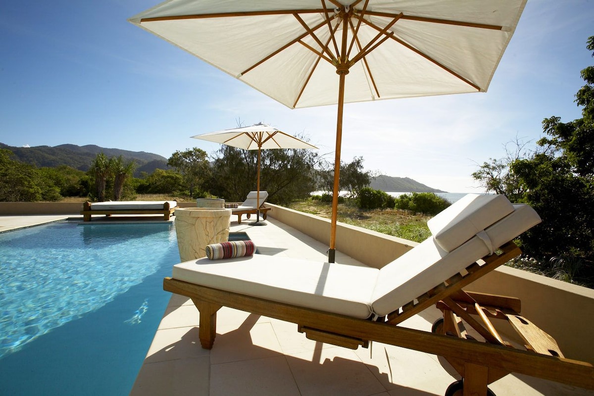 A serene poolside area is highlighted by wooden loungers, each accompanied by a large umbrella for shade. The tranquil water of the pool reflects the clear sky, while distant hills and lush greenery are visible in the background.