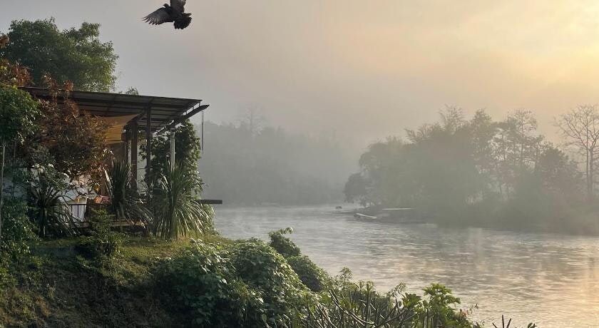The cottage is positioned along the tranquil banks of the River Kwai Noi, surrounded by lush greenery. Mist softly blankets the scene, enhancing the serene atmosphere. A bird is captured in mid-flight, adding a dynamic element to the peaceful riverside setting.