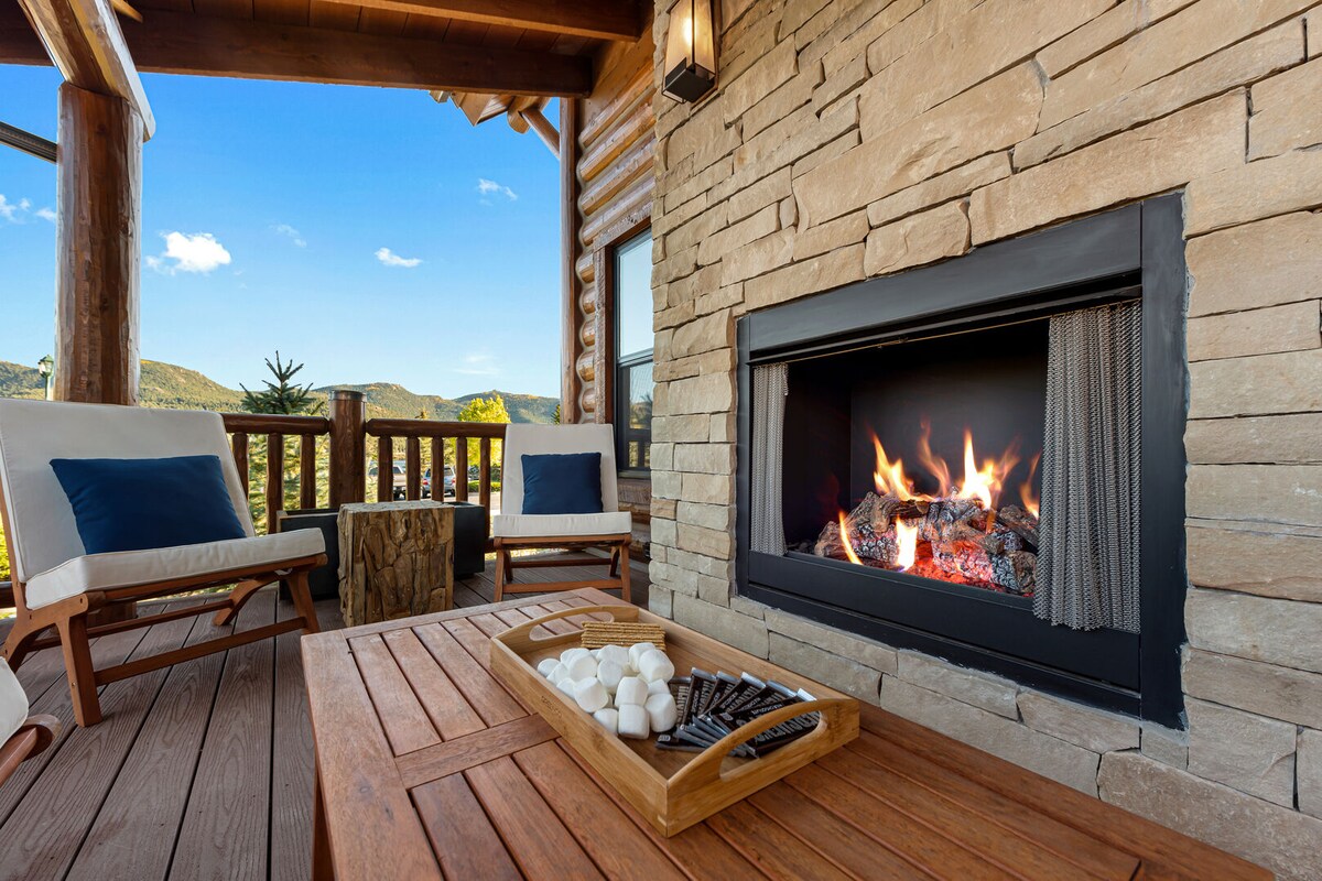 An outdoor seating area features two comfortable chairs positioned near a stone fireplace. A wooden table holds a tray with marshmallows and chocolate, ready for a cozy evening. Scenic mountain views are visible in the background, framed by the cabin's rustic wooden structure.