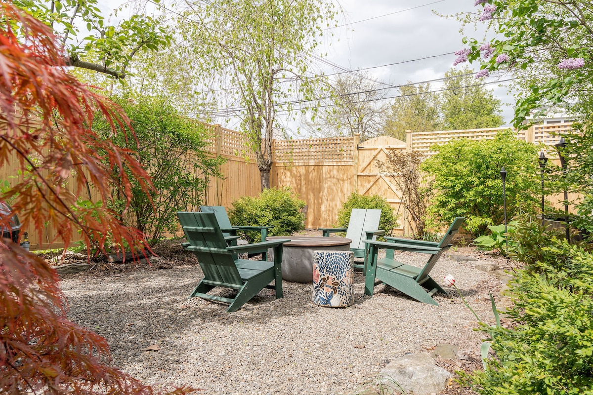 A serene outdoor space features four green Adirondack chairs arranged around a circular fire pit. Surrounding foliage includes shrubs and small trees, with a wood fence providing privacy. Natural textures are highlighted by gravel ground and stone accents, creating a tranquil retreat.