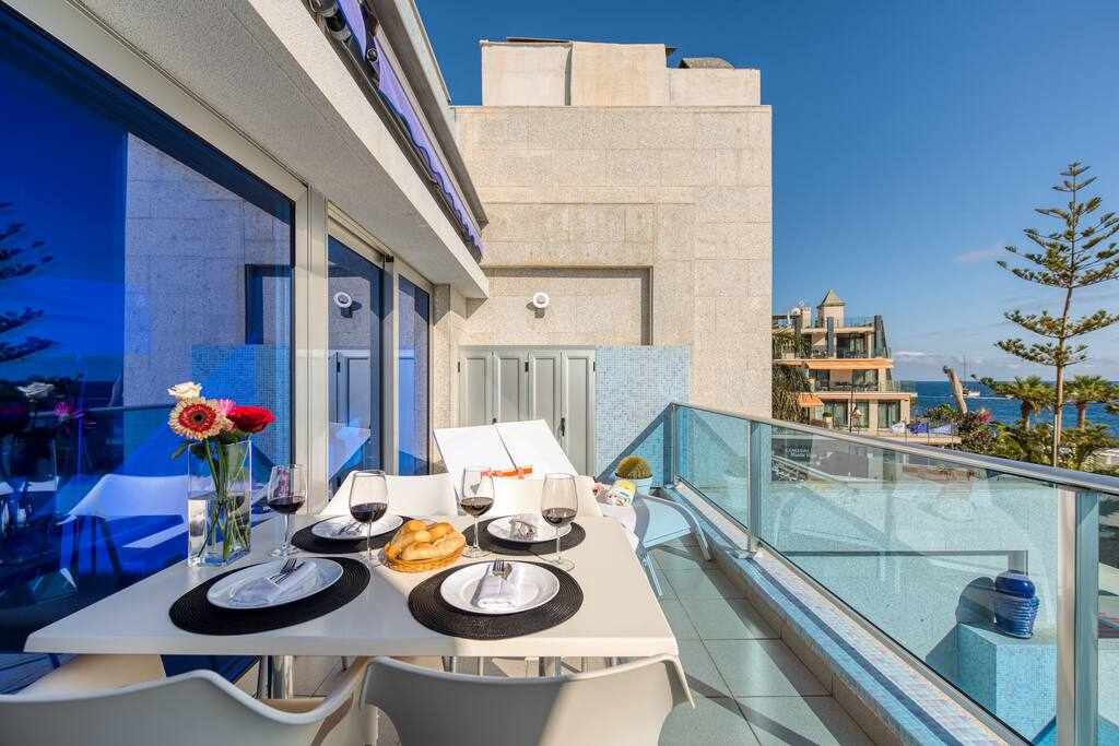 A modern terrace is shown, equipped with a dining table set for four. Vibrant flowers in a vase add color to the table, which is prepared with plates and glasses. The background features a clear blue sky, ocean views, and nearby buildings.