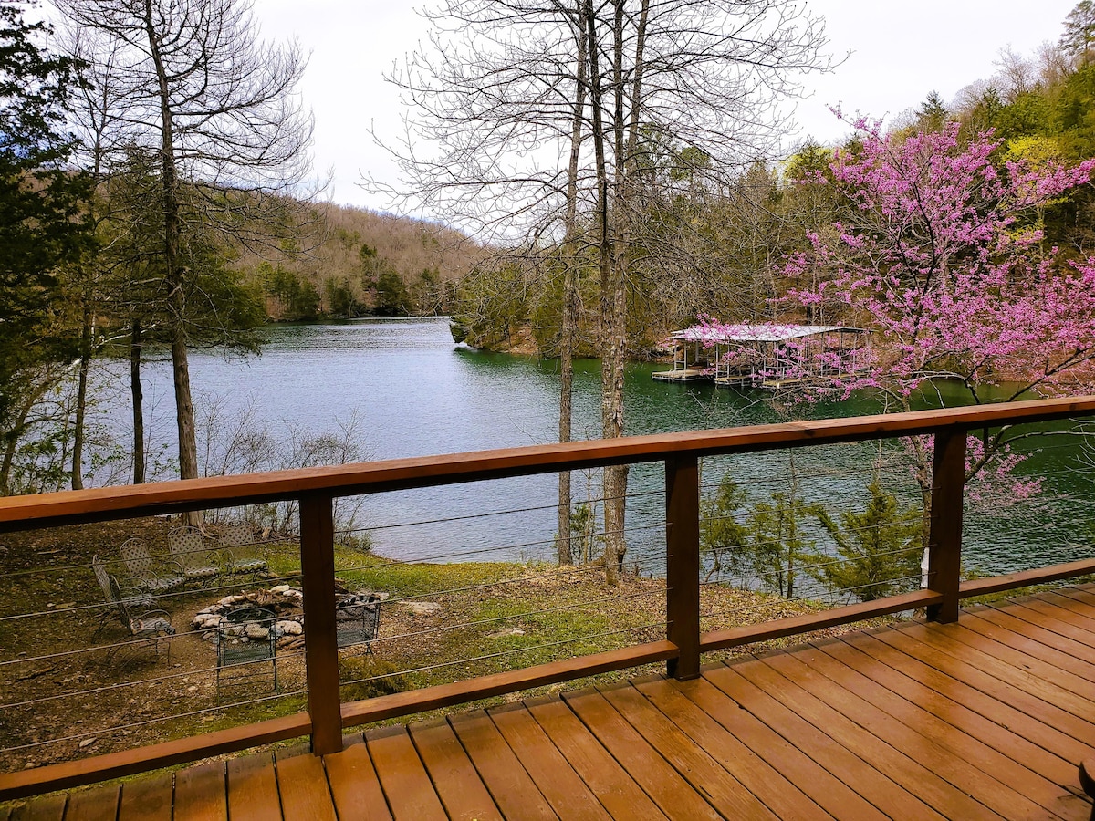 A scenic view of the lake is framed by the cabin's wooden deck. The water reflects the surrounding trees, with a noticeable pink flowering tree on the right. A fire pit area can be seen in the foreground, offering a serene outdoor gathering space.