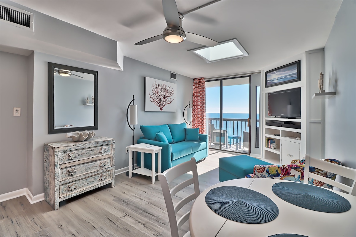 A bright living area showcases a turquoise sofa and a patterned lounge chair, complemented by light wood flooring. A glass balcony door offers expansive ocean views, while a round dining table with white chairs is positioned near the window. Decorative coastal art enhances the space.