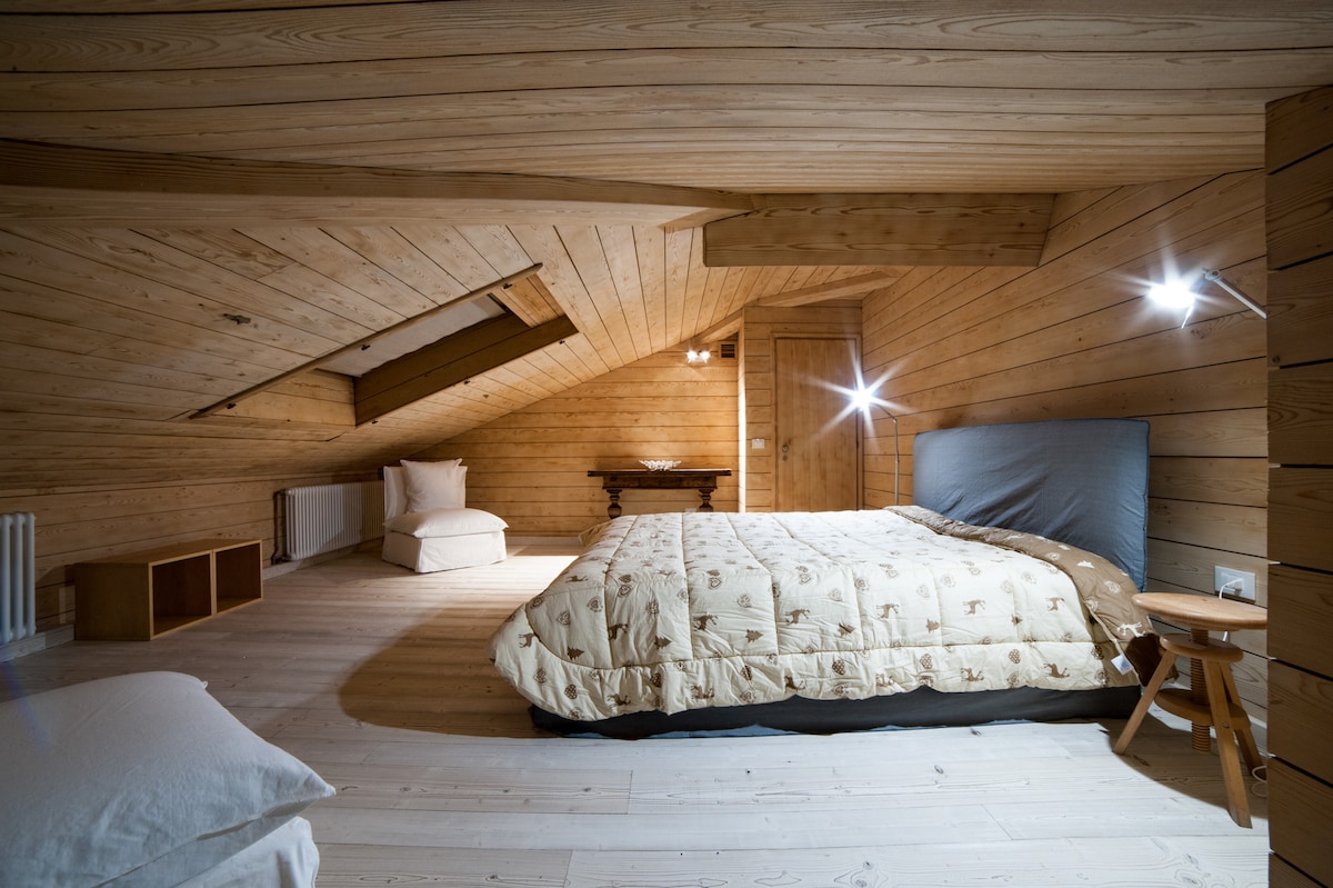 An attic bedroom features warm wooden walls and ceiling, creating a cozy atmosphere. A spacious bed is positioned centrally, covered with a beige comforter. A small bedside table stands beside the bed, while a chair and desk are visible in the background, providing functional space.