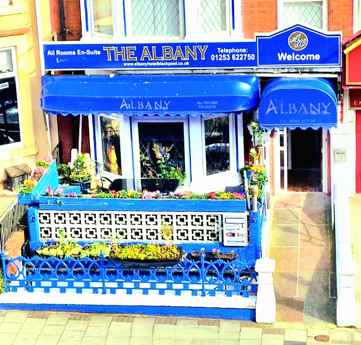 The exterior of The Albany Hotel showcases a vibrant blue facade adorned with floral arrangements. A welcoming entrance is framed by decorative railings, while a prominent sign displays the hotel's name. The overall appearance suggests a friendly and inviting atmosphere.