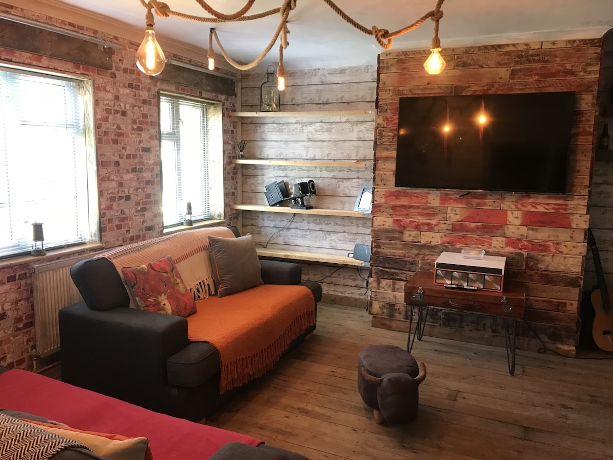 A lounge area features exposed brick walls and wooden shelves. A comfortable sofa is adorned with a colorful throw, while a TV is mounted on the wall. Lighting is provided by hanging bulbs, and a small stool complements the rustic decor.