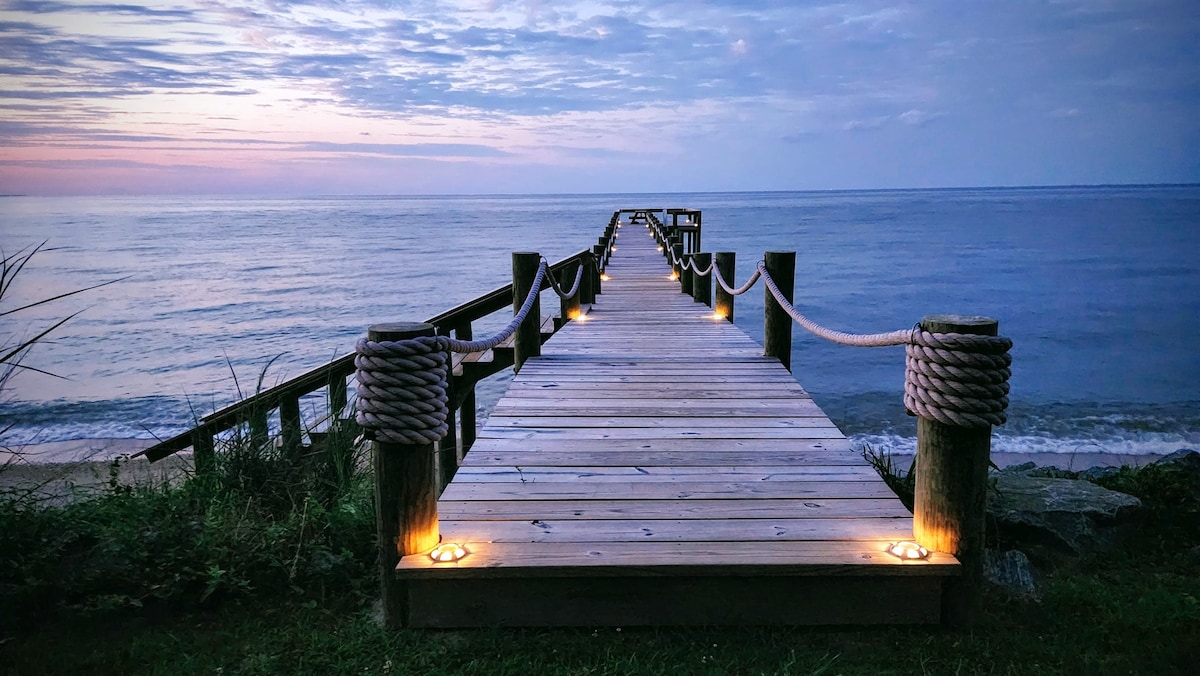 A wooden pier extending into calm waters is lined with soft lighting that enhances its structure. The surrounding landscape includes lush greenery leading to the shore. The sky reflects a blend of colors as day transitions to evening, adding a serene ambiance to the scene.