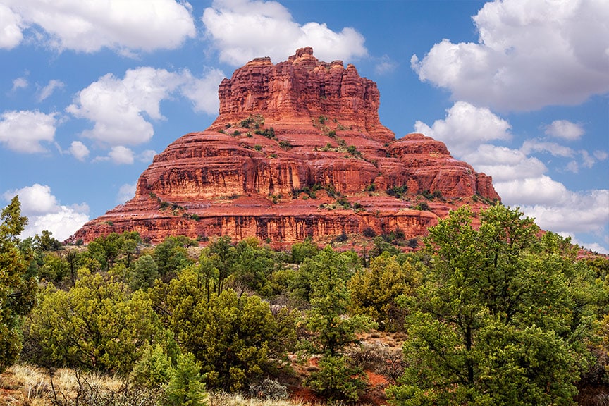 A striking red rock formation rises prominently against a blue sky dotted with fluffy clouds. The surrounding landscape features lush green foliage, creating a beautiful contrast with the warm tones of the rock. This scenic view captures the natural beauty of Sedona.