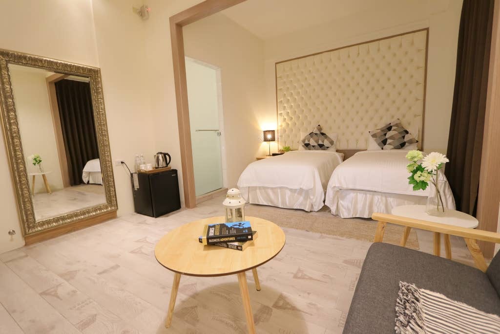 The room features two spacious beds with decorative headboards, complemented by soft lighting. A round coffee table is centrally located, surrounded by a cozy seating area with a gray sofa. A mirror adds depth to the space, and a small refrigerator and kettle are conveniently placed.