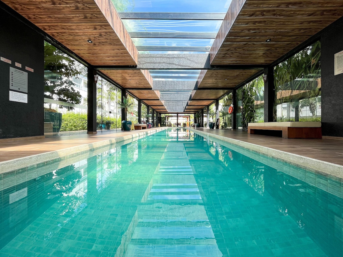 An indoor pool is featured with crystal-clear water and inviting reflections. A covered walkway runs alongside, lined with large windows that provide views of the surrounding greenery. The expansive wooden deck complements the tranquil ambiance.