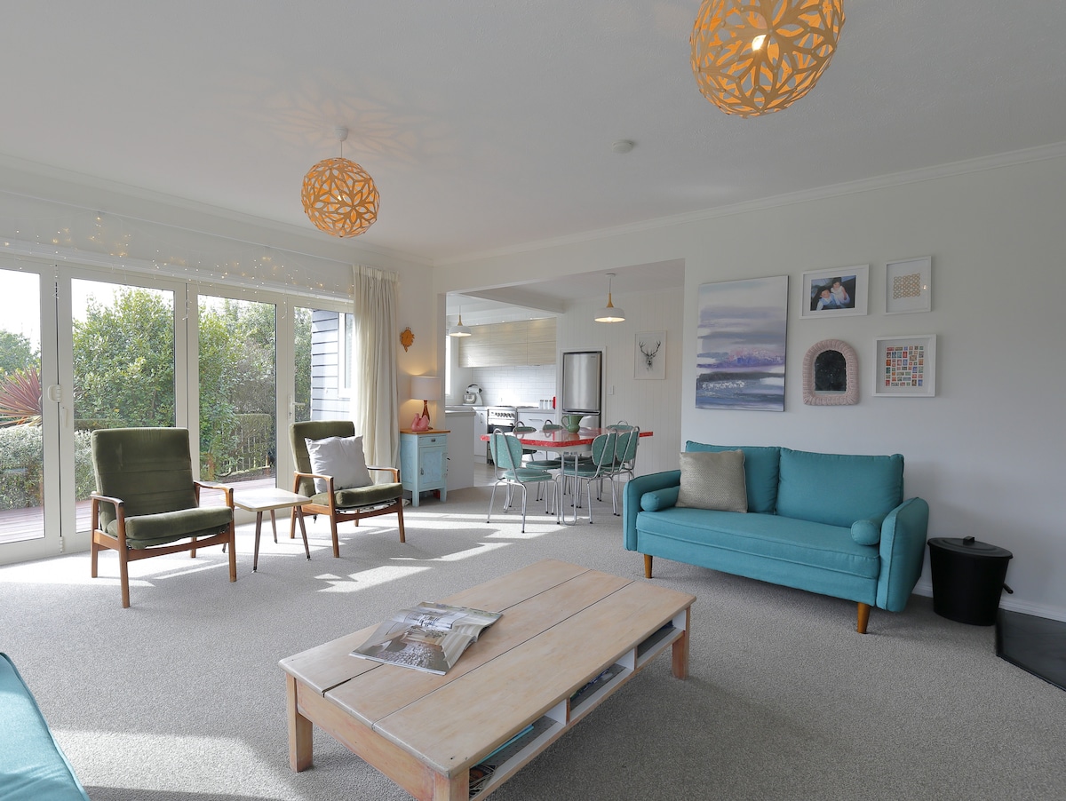 A well-lit living area features a soft blue couch and two vintage green armchairs, arranged around a wooden coffee table. Large glass doors open to a deck, with light fixtures adding a touch of warmth to the space. Vibrant artwork adorns the walls.