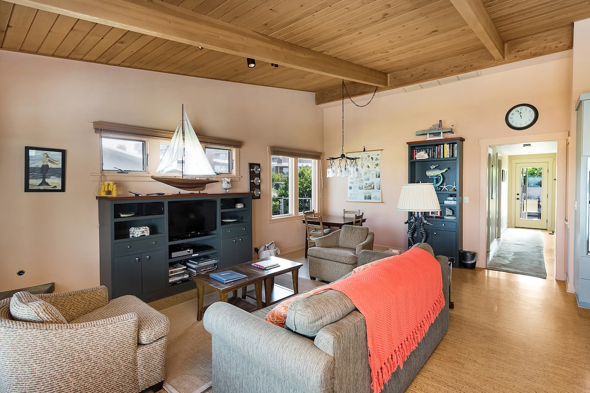 The living area features comfortable seating arrangements, including a spacious sofa adorned with a bright throw. Large windows allow natural light to fill the space, offering views of the surroundings. A light fixture hangs from the ceiling, while a hallway leads to other areas of the condo.