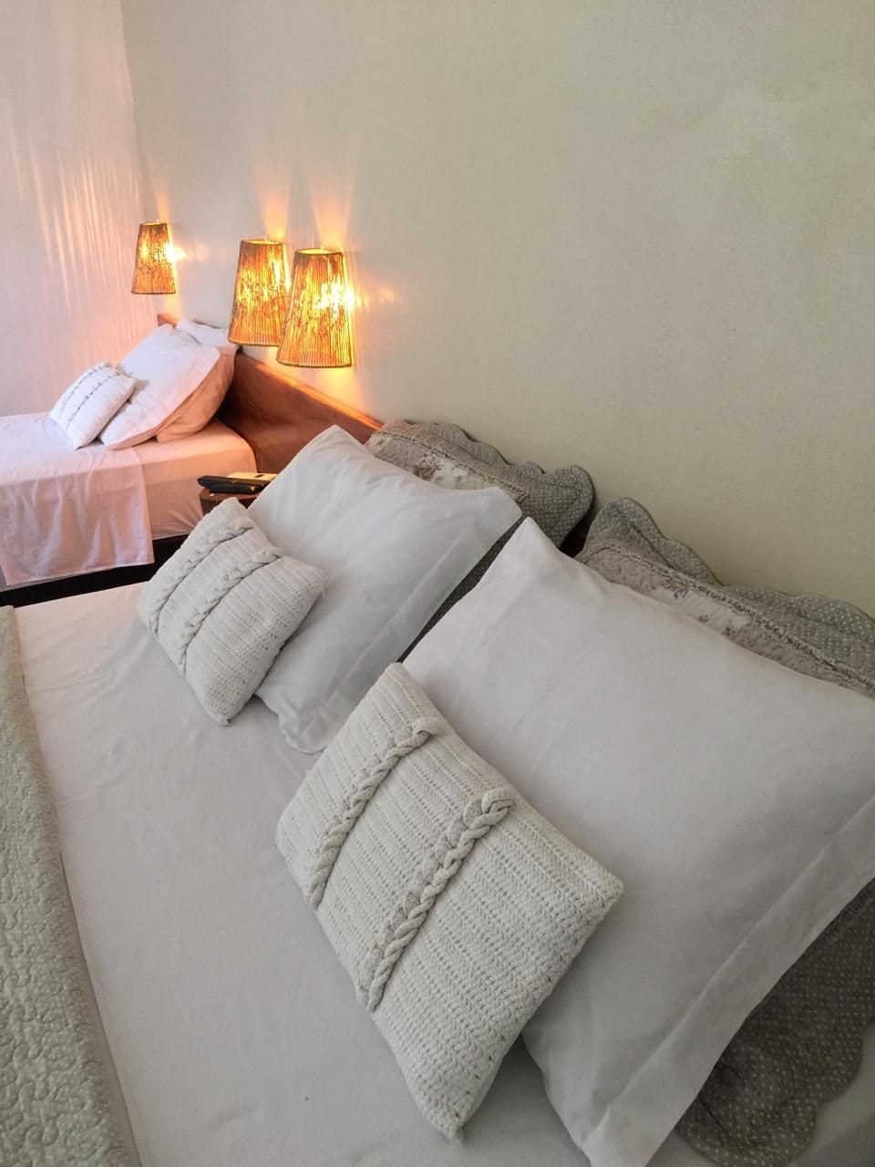 A comfortable sleeping area features a spacious bed with soft, white linens and decorative pillows. A smaller bed is positioned nearby. Ambient lighting is provided by two unique lamps, enhancing the cozy atmosphere in the room.