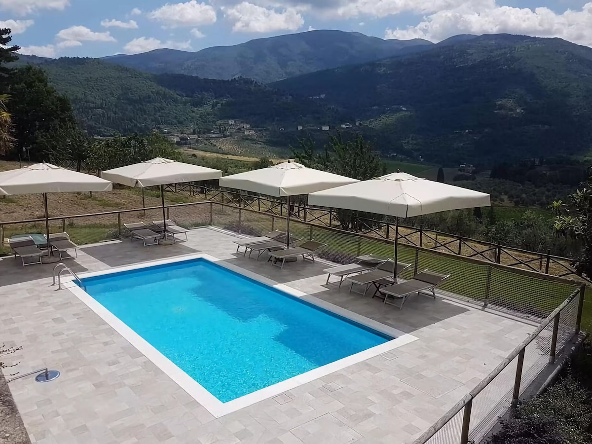 A well-maintained shared swimming pool is surrounded by comfortable lounge chairs and large umbrellas. The pool area offers a scenic view of the rolling hills and mountains, providing a relaxed outdoor space for guests to enjoy.