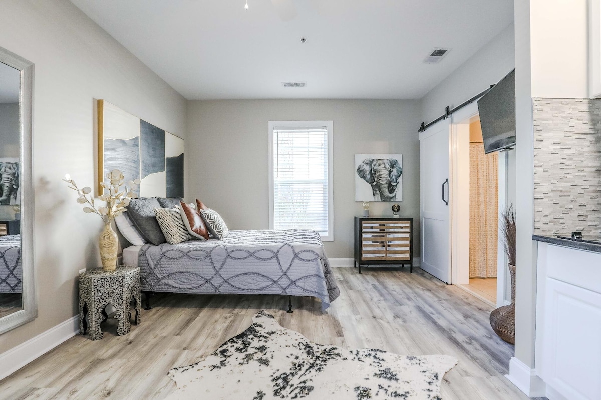 A spacious studio room is showcased, featuring a cozy bed with decorative pillows and a patterned blanket. A side table with a decorative vase adds a touch of elegance. Natural light filters through a window, illuminating the stylish decor that includes a large artwork of an elephant.
