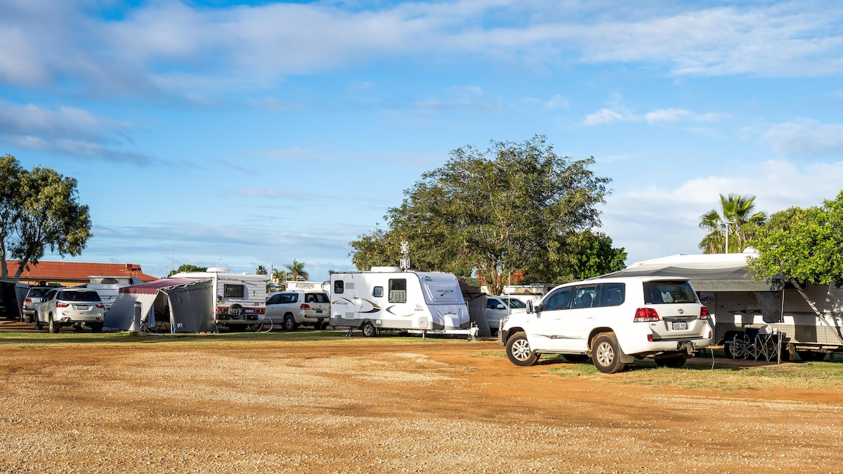A spacious caravan site is depicted, featuring several parked caravans and vehicles. The landscape includes a gravel surface with patches of grass and trees in the background, under a clear blue sky with scattered clouds.