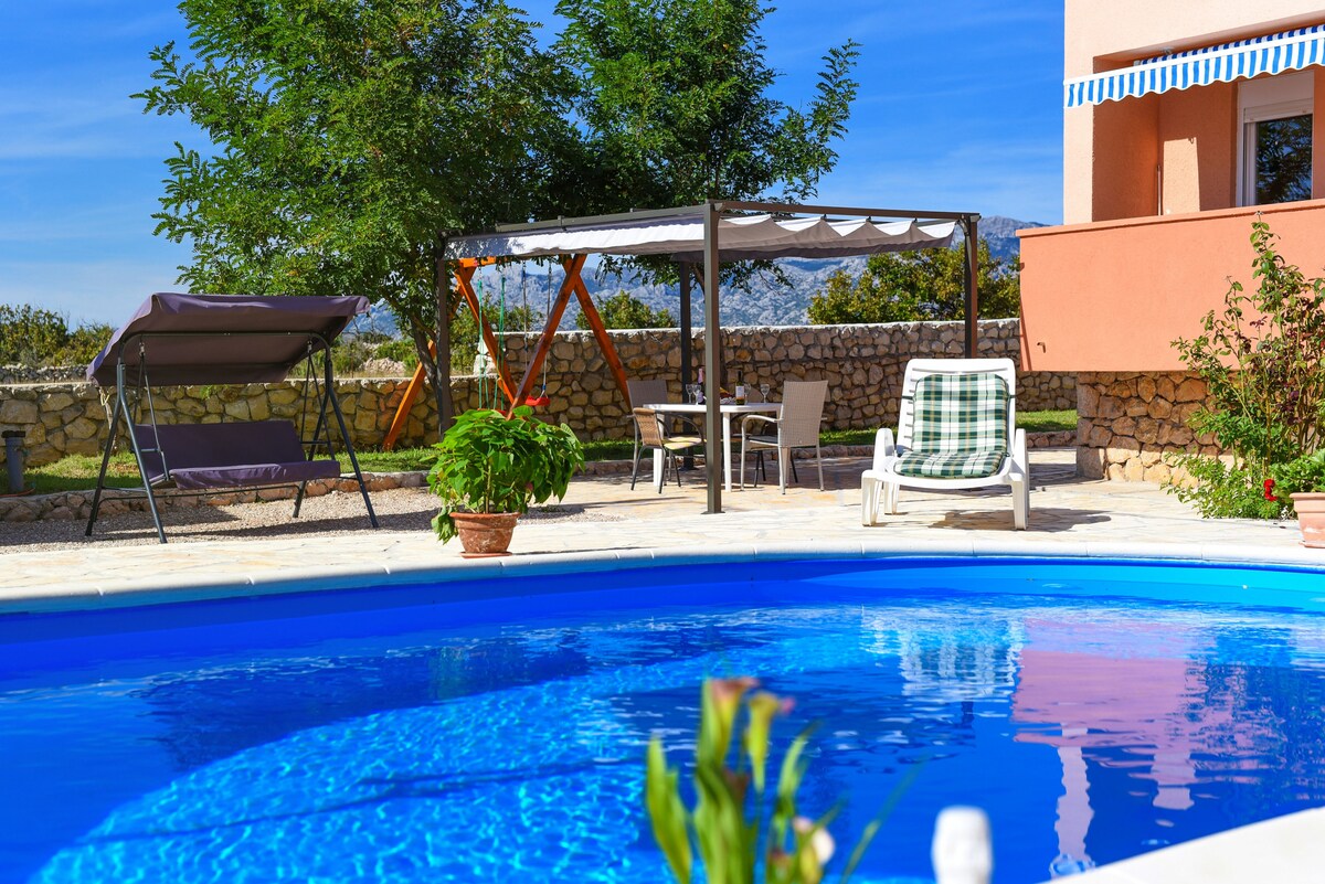 A crystal-clear swimming pool is surrounded by a sun terrace featuring loungers and a shaded gazebo. A dining area with a table is visible, complemented by a scenic landscape of greenery and distant hills.