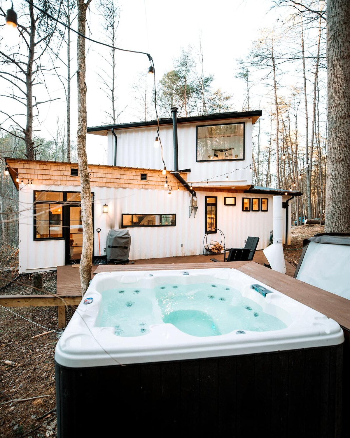 A hot tub is positioned on a large wooden deck, surrounded by trees. The two-story structure behind showcases large windows and inviting outdoor lighting. A grill and seating area are also visible, enhancing the tranquil outdoor setting.