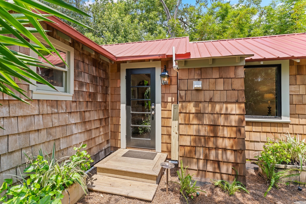 The entrance of the cottage features a wooden door framed by a welcoming porch. Natural wood siding and a red metal roof enhance the rustic charm. Lush greenery surrounds the entryway, providing a serene outdoor atmosphere.