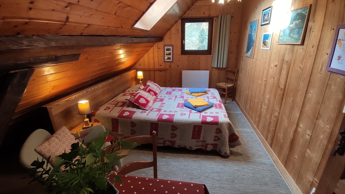 A cozy bedroom with a sloped wooden ceiling features a spacious bed adorned with patterned bedding and colorful towels. A comfortable chair complements a small table, while soft lighting is provided by bedside lamps. Artwork decorates the walls, enhancing the warm ambiance of the room.