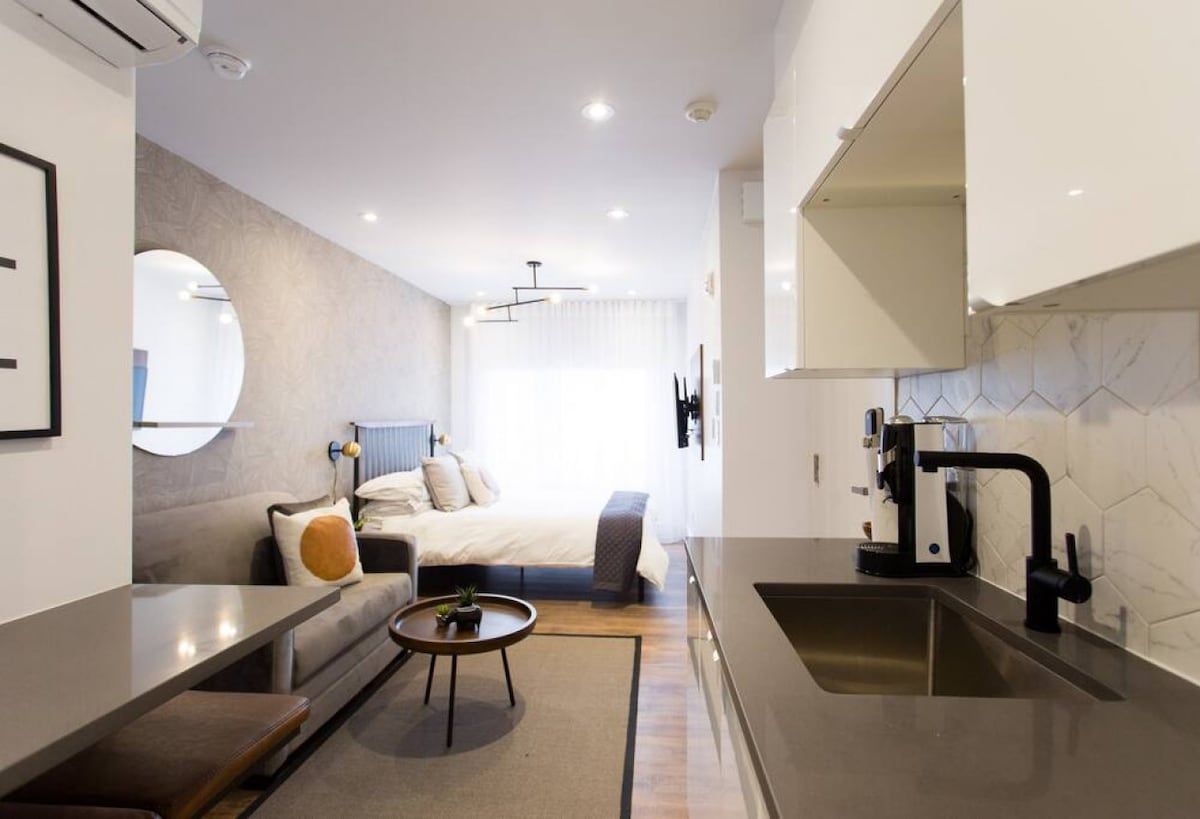 A modern studio layout is depicted, featuring a cozy sleeping area with a spacious bed framed by a light-colored wall. A round mirror complements the light-filled ambiance. A fully equipped kitchen is visible with sleek countertops and contemporary appliances, providing a functional living space.