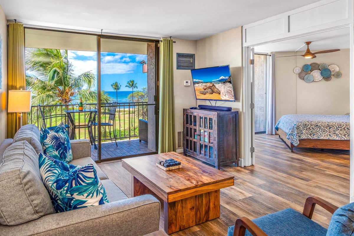 A spacious living area features a comfortable gray sofa adorned with tropical-patterned cushions. A wooden coffee table is centered in the room. Large sliding glass doors provide views of palm trees and the ocean, allowing natural light to enhance the inviting atmosphere. A flat-screen TV is mounted on the wall.