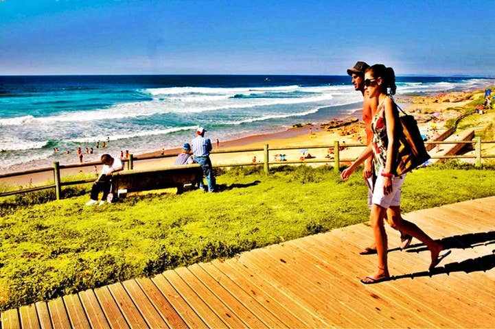 A sandy beach is visible in the background, with gentle waves lapping at the shore. Individuals can be seen enjoying the beach, while a wooden pathway leads towards the water. A grassy area is situated in the foreground, creating a welcoming space for relaxation.
