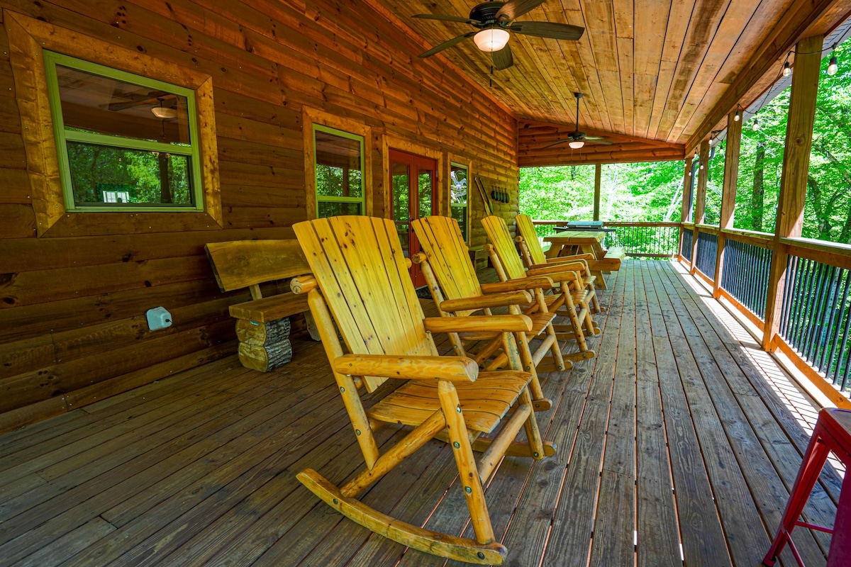 A spacious wrap-around porch is depicted, featuring several wooden rocking chairs arranged for relaxation. Natural light filters through large windows, highlighting the warm wood tones of the cabin. Lush greenery is visible outside, enhancing the tranquil outdoor setting.