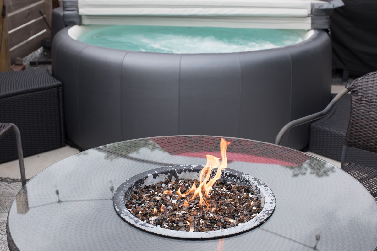 A Softub hot tub is visible in the background, surrounded by a wooden enclosure. In the foreground, a glass-top fire table features a visible flame, providing warmth. Cozy seating is arranged around the fire table for relaxation.