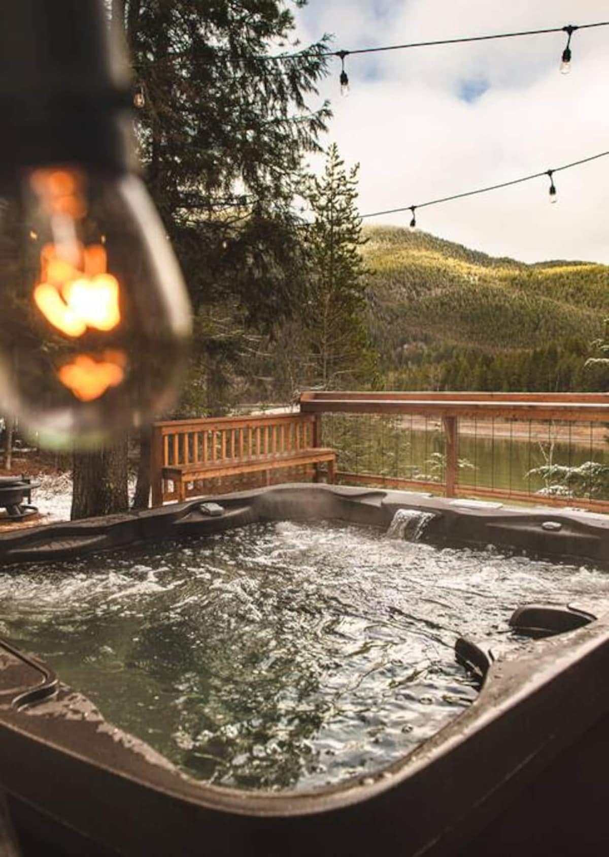 A hot tub, with steam rising from the water, is situated on a wooden deck overlooking a scenic landscape of trees and mountains. String lights overhead provide a warm glow, enhancing the serene atmosphere of the outdoor space.